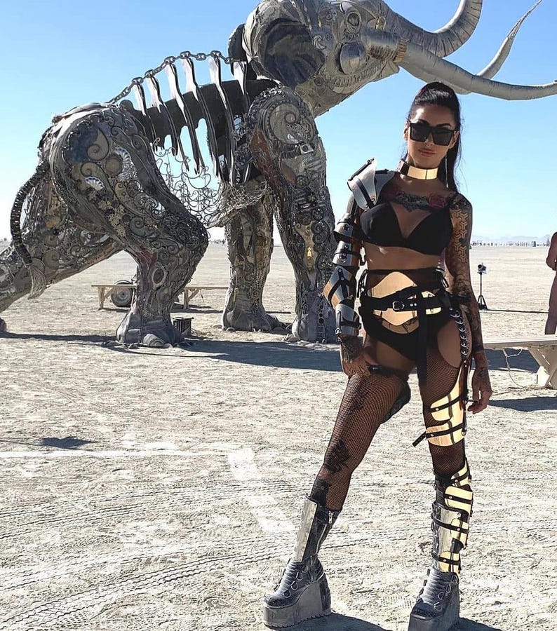 May include: A woman wearing a black and gold outfit with a metal harness and fishnet stockings. She is standing in front of a large metal sculpture of an elephant.