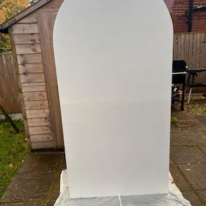 May include: A large, white, arched backdrop stands outdoors on a stone patio. The backdrop is made of two rectangular panels joined together. The top is rounded, creating an arch shape. The backdrop is resting on a white sheet.