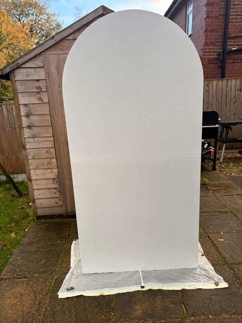 May include: A large, white, arched backdrop stands outdoors on a stone patio. The backdrop is made of two rectangular panels joined together. The top is rounded, creating an arch shape. The backdrop is resting on a white sheet.