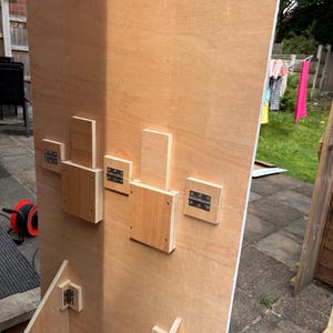 May include: A partially constructed wooden structure with light brown plywood panels. Several small, rectangular wooden blocks are attached to the plywood with metal hinges. The structure appears to be outdoors, with a glimpse of a clothesline in the background.