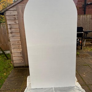 May include: A large, white, arched backdrop stands outdoors on a stone patio. The backdrop is made of two rectangular panels joined together. The top is rounded, creating an arch shape. The backdrop is resting on a white sheet.