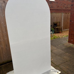 May include: A large, white, arched wooden panel, likely for event decor. The panel is rectangular with a rounded top, and is resting on a protective sheet outdoors. The background includes a wooden fence, brick building, and a window.