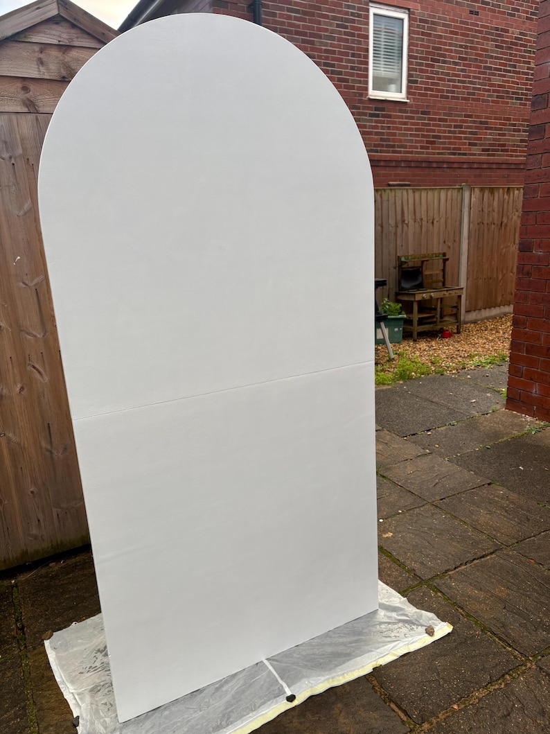 May include: A large, white, arched wooden panel, likely for event decor. The panel is rectangular with a rounded top, and is resting on a protective sheet outdoors. The background includes a wooden fence, brick building, and a window.
