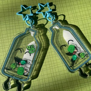 Pode incluir: Dois chaveiros de acrílico transparente em forma de garrafa, cada um com contorno azul e topo em forma de estrela. Dentro de cada garrafa há personagens de desenhos animados verdes e um fundo branco. Os chaveiros estão em uma superfície verde texturizada.