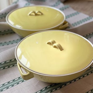 Può includere: Due ciotole da portata in ceramica gialla con coperchi. Le ciotole hanno un bordo bianco e un design semplice con due manici su ogni lato.