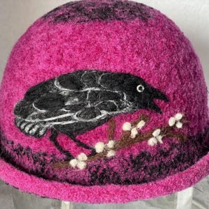 May include: A vibrant magenta felted wool hat featuring a needle-felted black crow perched on a brown branch with small white blossoms. The hat has a rounded crown and a short, turned-up brim, with dark purple accents along the base.