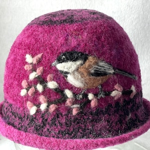 May include: A magenta felted cloche hat featuring a needle-felted chickadee perched on a branch with small pink and white buds. The hat has a dark mottled texture around the base and crown, creating a distinct contrast against the bright magenta.