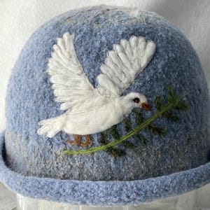 May include: A light blue felted hat featuring a white dove in flight with its wings spread wide. The dove holds a thin green olive branch in its beak. The hat has a rounded crown and a rolled brim, showcasing a textured, wool-like surface.