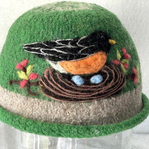 May include: A green felted hat featuring a needle-felted design of a black and orange bird sitting in a brown nest with two light blue eggs. The hat has a beige band around the base and decorative branches with small red berries on the sides.