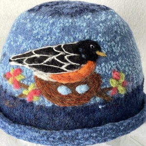 May include: A blue felted hat featuring a needle-felted design of a black and orange bird sitting in a brown nest with two light blue eggs. The hat includes small pink and green floral accents on branches surrounding the nest.