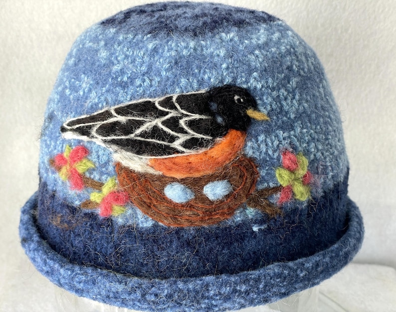 May include: A blue felted hat featuring a needle-felted design of a black and orange bird sitting in a brown nest with two light blue eggs. The hat includes small pink and green floral accents on branches surrounding the nest.