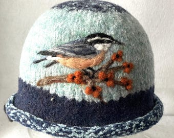 Pode incluir: Um chapéu de feltro com base azul clara e aba azul escura. O chapéu apresenta um design de feltro com agulha de um pequeno pássaro com plumagem preta, branca e laranja pousado num ramo castanho com pequenas bagas cor de laranja.