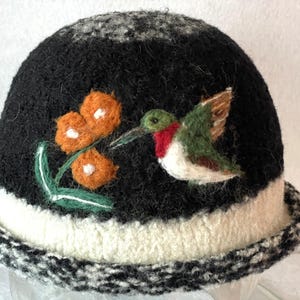 May include: A black felted hat featuring a needle-felted design of a green and red hummingbird hovering near three orange flowers with white centers. The hat has a wide white band above a black and white speckled brim with a textured finish.
