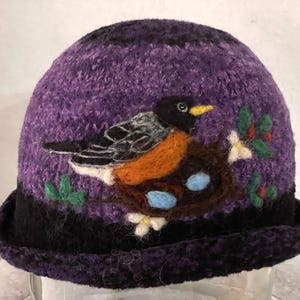 May include: A purple felted hat featuring a needle-felted design of a robin perched on a brown nest containing two light blue eggs. The bird has a black head, orange chest, and grey wings. Green leaves and white flowers accent the nest.