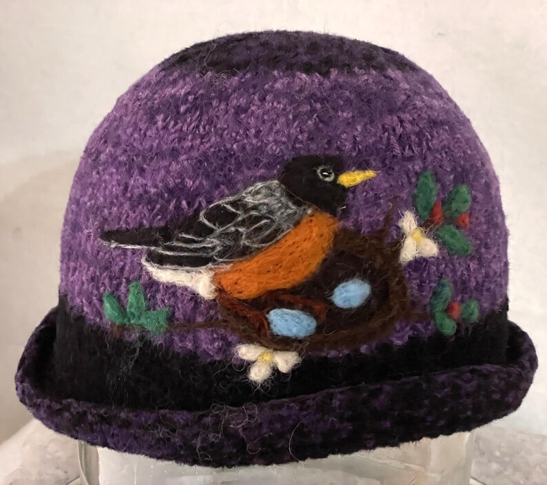 May include: A purple felted hat featuring a needle-felted design of a robin perched on a brown nest containing two light blue eggs. The bird has a black head, orange chest, and grey wings. Green leaves and white flowers accent the nest.