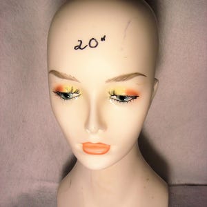 May include: A smooth, beige mannequin head featuring painted facial details including orange and yellow eyeshadow, long black eyelashes, and orange lips. The text 20" is written in black ink on the forehead of the mannequin head.