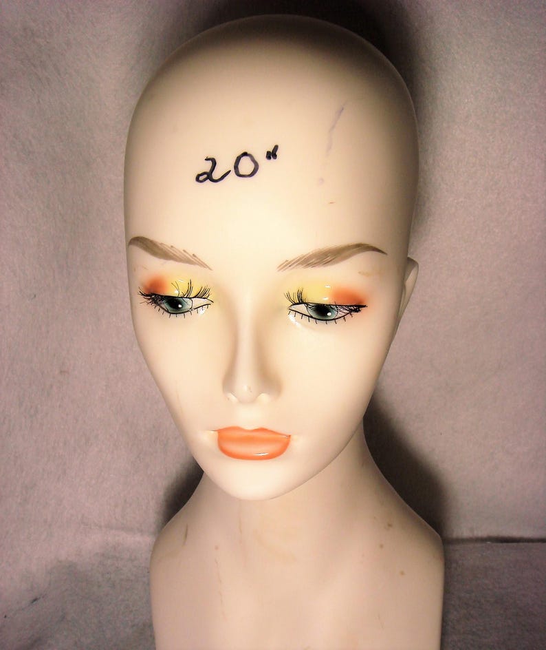 May include: A smooth, beige mannequin head featuring painted facial details including orange and yellow eyeshadow, long black eyelashes, and orange lips. The text 20" is written in black ink on the forehead of the mannequin head.