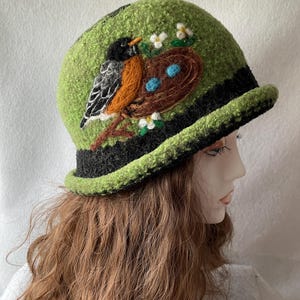 May include: A lime green felted cloche hat featuring a black brim and a needle-felted design of a robin perched on a branch next to a brown nest containing two light blue eggs, accented with small white flowers and green leaves.
