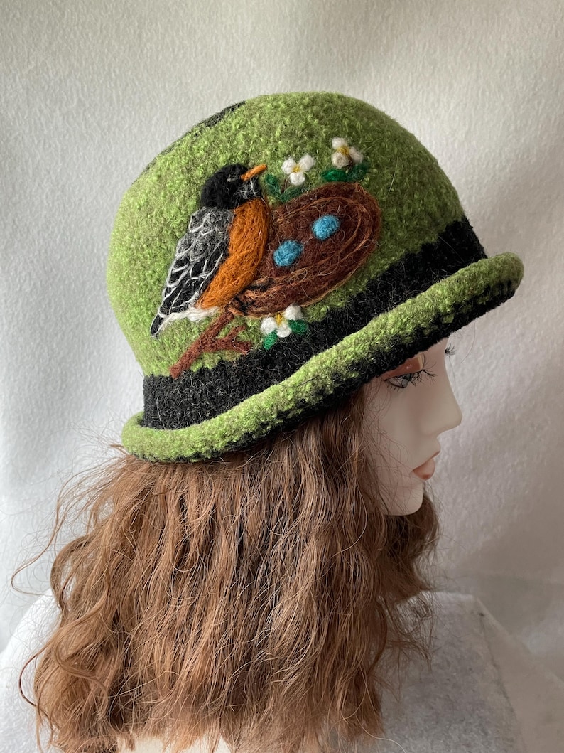 May include: A lime green felted cloche hat featuring a black brim and a needle-felted design of a robin perched on a branch next to a brown nest containing two light blue eggs, accented with small white flowers and green leaves.