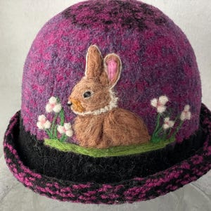 May include: A felted hat in shades of purple and magenta featuring a needle-felted brown rabbit sitting on a green patch of grass. The hat has a black brim with a magenta pattern and small white flowers surrounding the rabbit.
