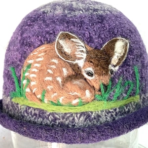 May include: A purple felted hat featuring a needle-felted design of a brown fawn with white spots curled up in green grass. The hat has a textured, mottled purple surface and a rolled brim, showcasing detailed fiber art craftsmanship.