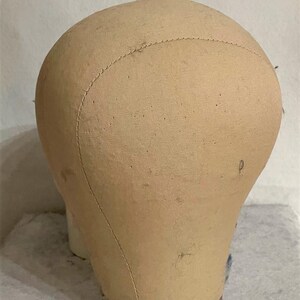 May include: A beige canvas-covered mannequin head with a rounded top and a smooth, fabric-wrapped surface. The head features visible stitched seams running vertically across the crown and a circular, ribbed base in a muted reddish-brown tone.