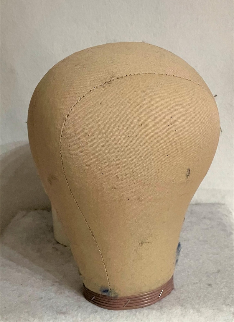 May include: A beige canvas-covered mannequin head with a rounded top and a smooth, fabric-wrapped surface. The head features visible stitched seams running vertically across the crown and a circular, ribbed base in a muted reddish-brown tone.