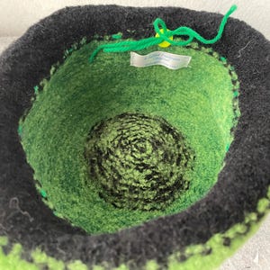 May include: A bowl-shaped felted container featuring a black outer rim and a vibrant green interior with a dark green and black spiral pattern at the base. A bright green yarn tie is attached to a small yellow button, and a white label reads A Felted Fantasy Original.