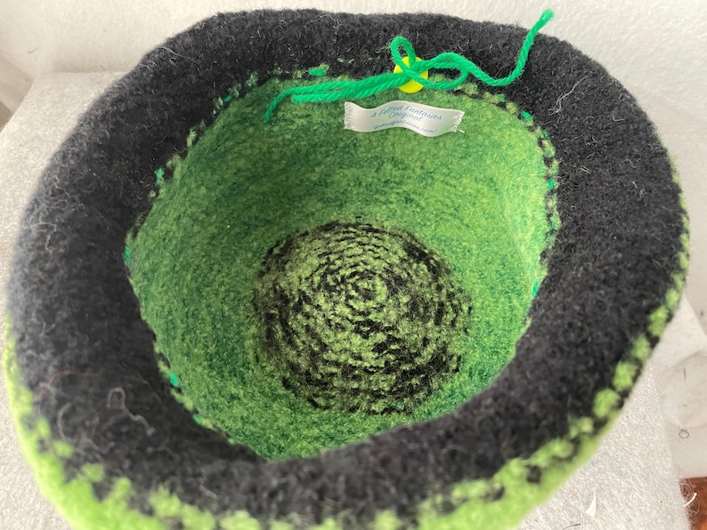 May include: A bowl-shaped felted container featuring a black outer rim and a vibrant green interior with a dark green and black spiral pattern at the base. A bright green yarn tie is attached to a small yellow button, and a white label reads A Felted Fantasy Original.