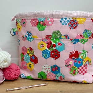 May include: Pink fabric project bag with a patchwork design of hexagons in various colours and patterns. The bag has a drawstring closure and a handle.