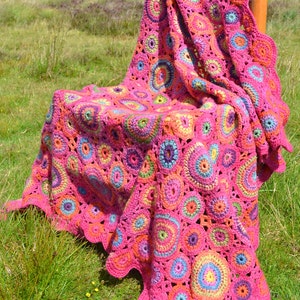May include: A pink crocheted throw blanket with a colorful circular pattern. The blanket is draped over a wooden chair and is sitting on a grassy field.