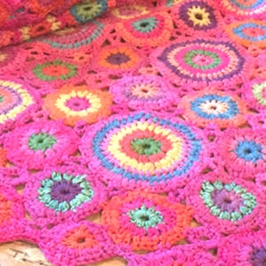 May include: A crocheted blanket with a pink background and colorful circular patterns. The blanket is made with a variety of colors, including pink, red, orange, yellow, green, blue, and purple.