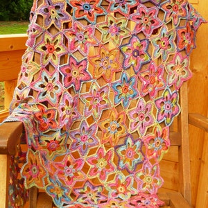 Puede incluir: Un chal de ganchillo colorido con un patrón floral. El chal está hecho con una variedad de colores brillantes, incluyendo rosa, morado, azul, verde y amarillo. El chal está drapeado sobre una silla de madera.