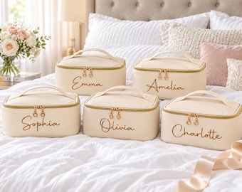 May include: Five rectangular cream-colored cosmetic bags with gold-tone zippers and handles. Each bag features a unique name embroidered in gold script: Emma, Amelia, Sophia, Olivia, and Charlotte. The bags are arranged on a white bedspread.