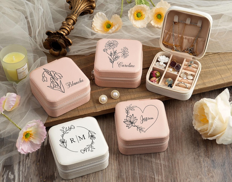 May include: Set of four pink and white jewelry boxes with floral designs and personalized names. The boxes are square and have a hinged lid. Each box has a different floral design and name: Blanche, Caroline, Jessica, and R/M.
