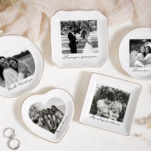 Op de afbeelding: Een verzameling witte keramische sieradenschaaltjes in verschillende vormen, elk met een zwart-witfoto en gouden rand. De schaaltjes omvatten ronde, vierkante en hartvormige ontwerpen, met opschriften als "Best Wishes" en "Love you mom".