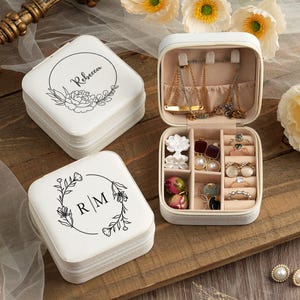 May include: Two white square jewelry boxes with floral designs and personalized names. The top box is open and has compartments for rings, earrings, and necklaces. The bottom box has the initials "R/M" on the lid.