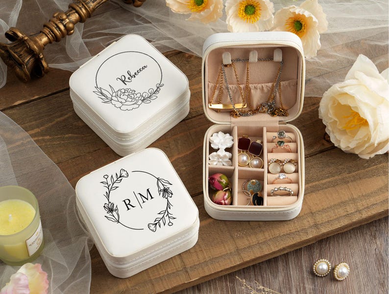 May include: Two white square jewelry boxes with floral designs and personalized names. The top box is open and has compartments for rings, earrings, and necklaces. The bottom box has the initials "R/M" on the lid.