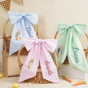 May include: Three Easter baskets with large seersucker bows in blue, pink, and green. Each bow has a name embroidered on it, along with an embroidered bunny design. The baskets are filled with Easter grass.