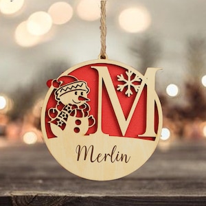 以下が含まれることがあります： 雪だるまと文字「M」がくり抜かれた木製のクリスマスオーナメント。オーナメントは赤い背景で、「Merlin」という名前が筆記体で書かれています。「M」の隣には雪の結晶がくり抜かれています。