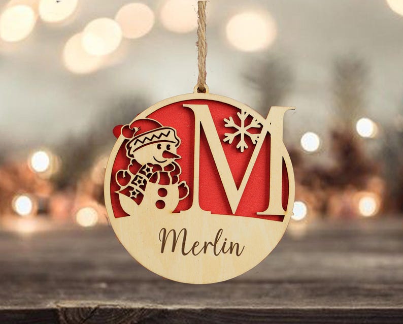 以下が含まれることがあります： 雪だるまと文字「M」がくり抜かれた木製のクリスマスオーナメント。オーナメントは赤い背景で、「Merlin」という名前が筆記体で書かれています。「M」の隣には雪の結晶がくり抜かれています。