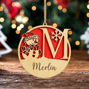 以下が含まれることがあります： 雪だるまと切り抜かれた文字「M」が付いた木製のクリスマスオーナメント。オーナメントは赤い背景で、「Merlin」という名前が筆記体で書かれています。吊り下げ用の紐が付いています。