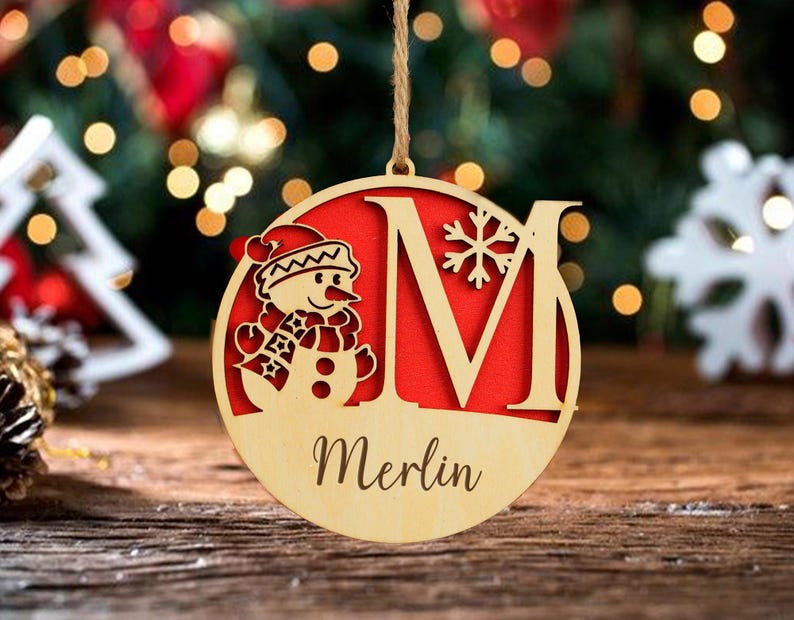 以下が含まれることがあります： 雪だるまと切り抜かれた文字「M」が付いた木製のクリスマスオーナメント。オーナメントは赤い背景で、「Merlin」という名前が筆記体で書かれています。吊り下げ用の紐が付いています。