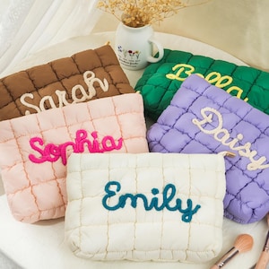 Op de afbeelding: Een verzameling gewatteerde make-up tassen in bruin, roze, groen, paars en wit. Elke tas heeft een geborduurde naam: Sarah, Sophia, Bella, Daisy en Emily. De tassen liggen op een witte ondergrond, met make-up kwasten.