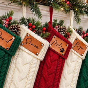 Puede incluir: Cinco calcetines navideños de punto trenzado en colores alternos verde, blanco y rojo. Cada calcetín tiene un parche rectangular de cuero color canela sujeto con cuatro remaches metálicos, grabado con los nombres David, Simon, Milo con una huella, Rigon y Frank.