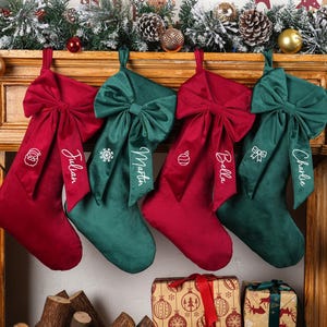 May include: Four Christmas stockings in red and green velvet hang from a wooden mantel. Each stocking has a large bow and is embroidered with a name. Festive decorations, including pinecones and ornaments, adorn the mantel.