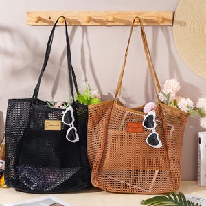 Könnte beinhalten: Zwei Netztaschen, eine schwarze und eine braune, jeweils mit langen Trägern. Die schwarze Tasche hat ein goldenes Etikett mit der Aufschrift "Divana" und die braune Tasche ein braunes Etikett. Beide Taschen haben herzförmige Sonnenbrillen und sind mit Blumen gefüllt.