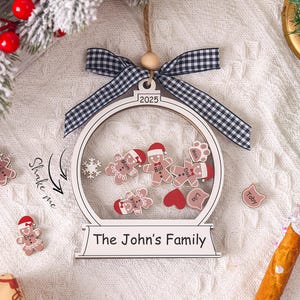 May include: A white wooden Christmas ornament shaped like a snow globe, with a black and white checkered bow. Inside are gingerbread people and hearts. The text reads "The John's Family" and "2025".