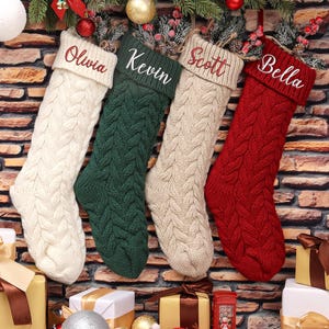 Puede incluir: Cuatro calcetines de Navidad personalizados con los nombres "Olivia", "Kevin", "Scott" y "Bella" bordados en ellos. Los calcetines están hechos de tejido de punto con un patrón de punto de cable. Los calcetines están colgados en una pared de ladrillo con decoraciones navideñas.