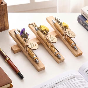 May include: Wooden pens with silver detailing and personalised names are displayed on wooden stands, decorated with dried flowers and tags. A leather-bound notebook and a pen are also visible, along with open books and pencils.